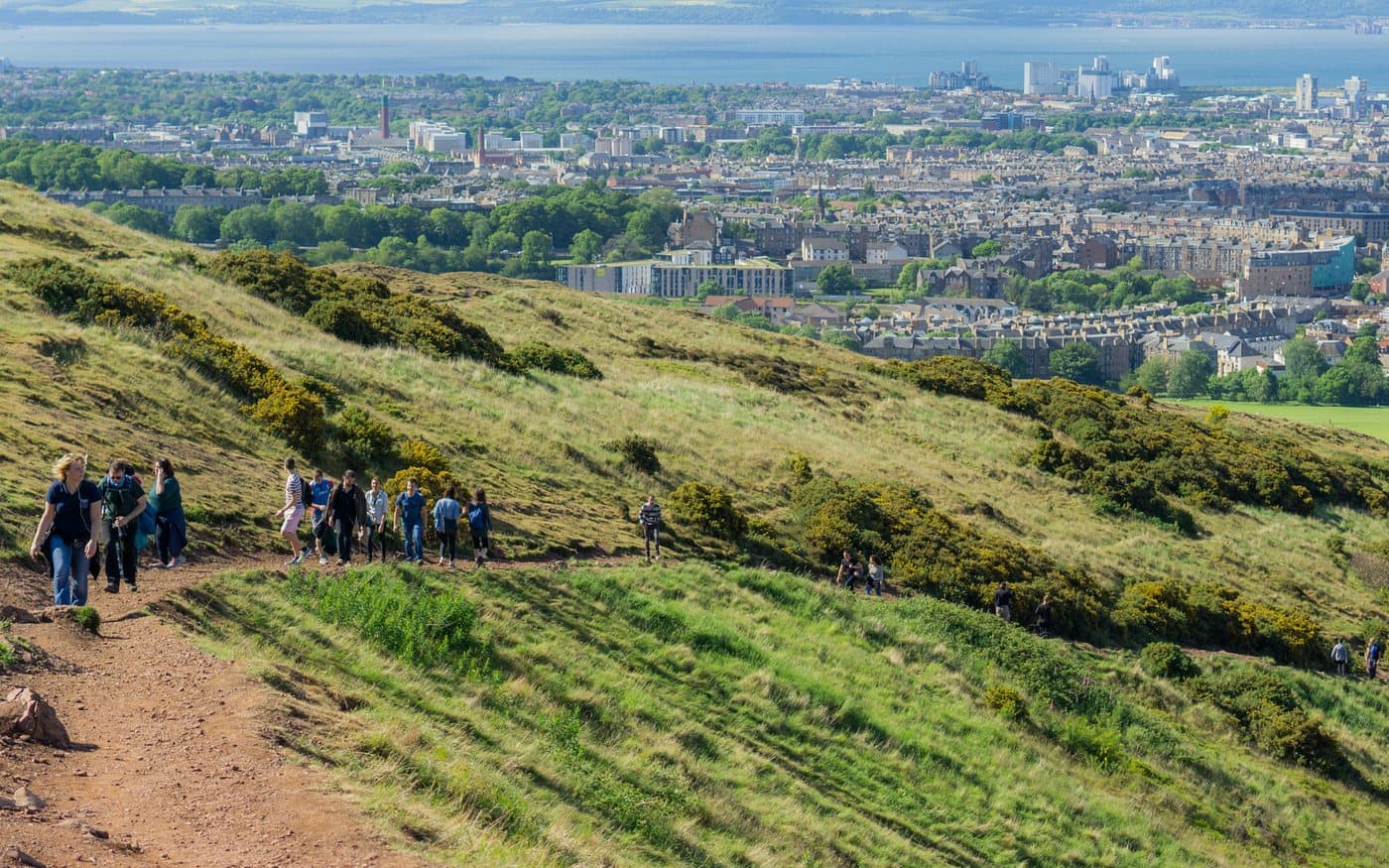 Walkers in ordinary clothes strung out along a broad eroded path on the grassy flank of Arthur's Seat, with Edinburgh's tenements and the Firth of Forth behind