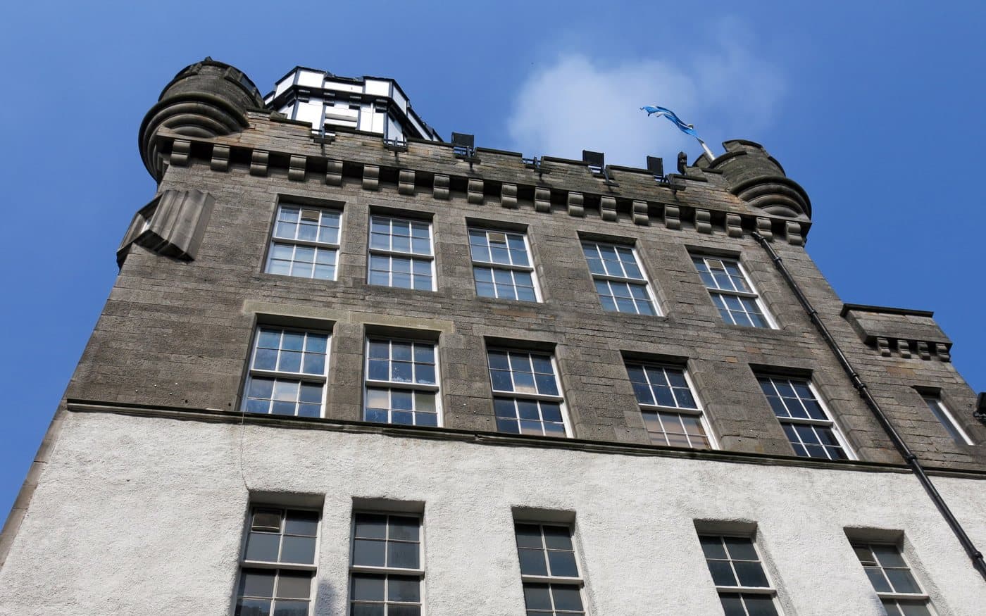 Looking straight up the facade of the Outlook Tower from Castlehill, showing a white harled lower building with taller grey stone floors above it and the glazed camera obscura lantern at the top
