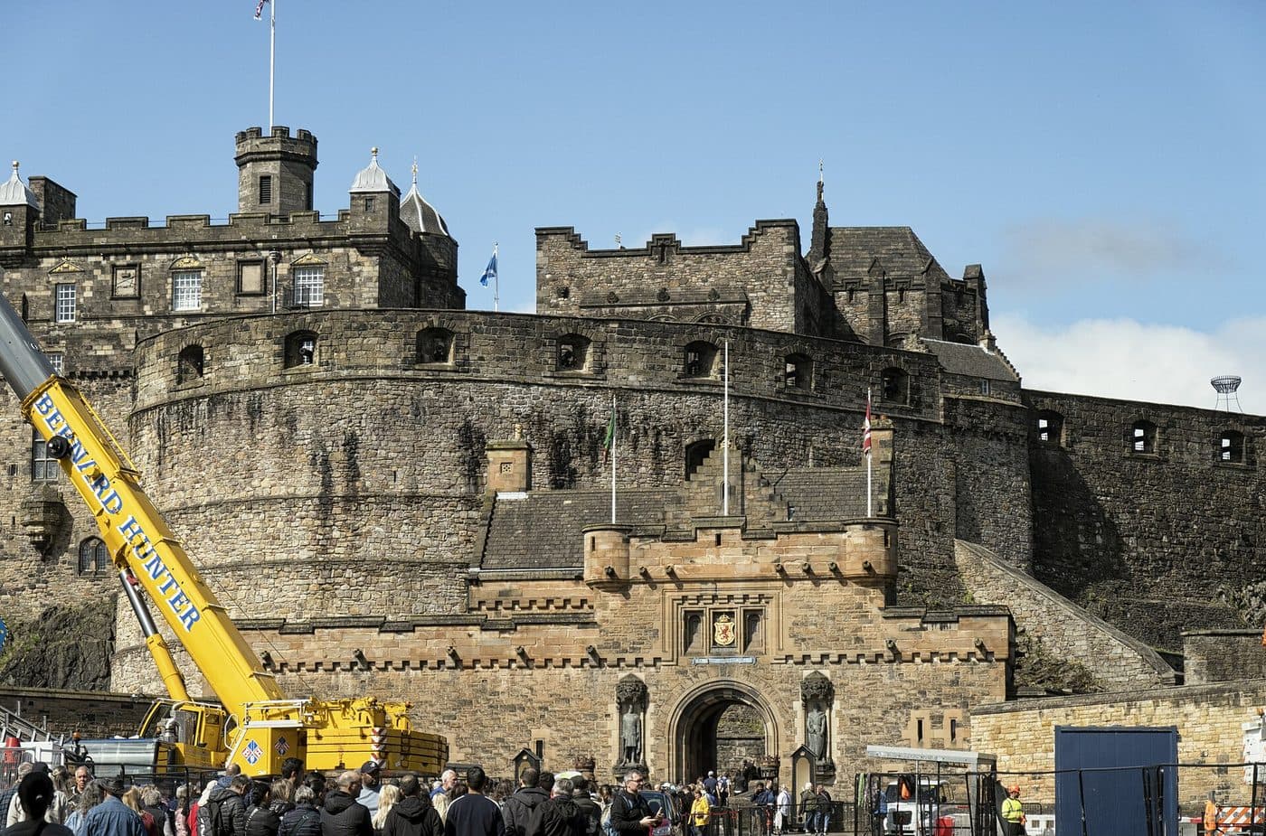 Crowds gathered at the Edinburgh Castle gatehouse below the statues of Wallace and Bruce, with a yellow mobile crane and construction hoarding on the esplanade