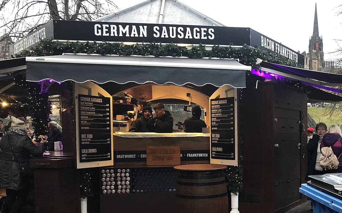 A German sausage chalet at the Edinburgh Christmas Market in East Princes Street Gardens, its price boards facing the walkway