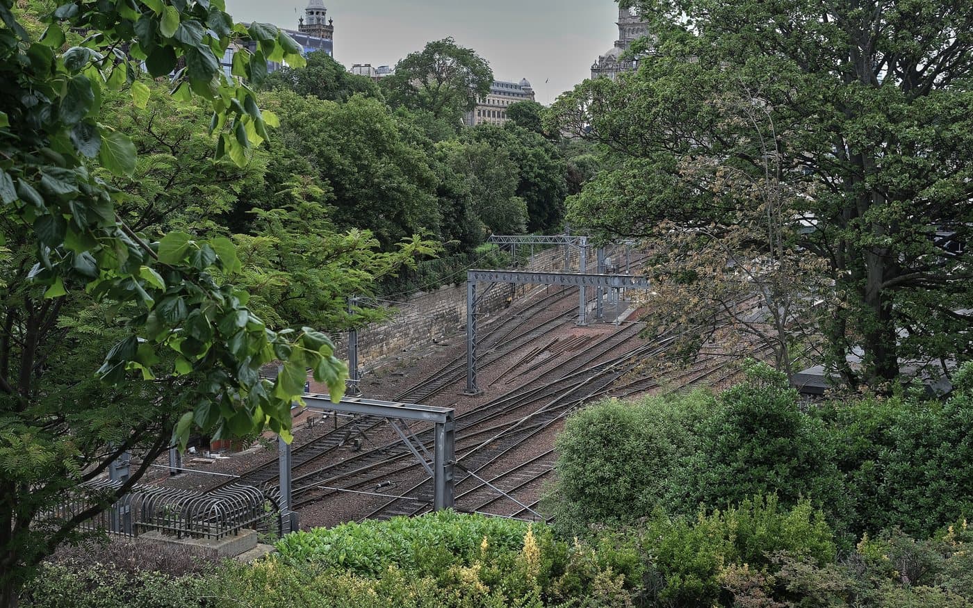 The approach tracks into Edinburgh Waverley seen from above, running through a deep green cutting below the city, with rooftops and a clock tower on the skyline