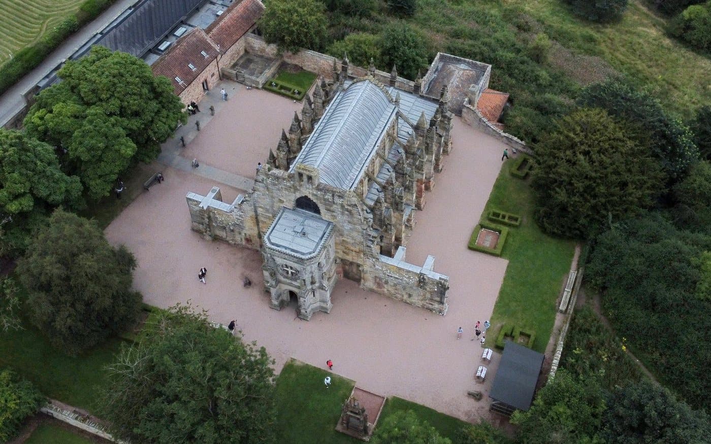 Rosslyn Chapel seen from directly above, a compact cross-shaped building with a lead roof surrounded by a gravel apron, clipped hedges and a handful of visitors