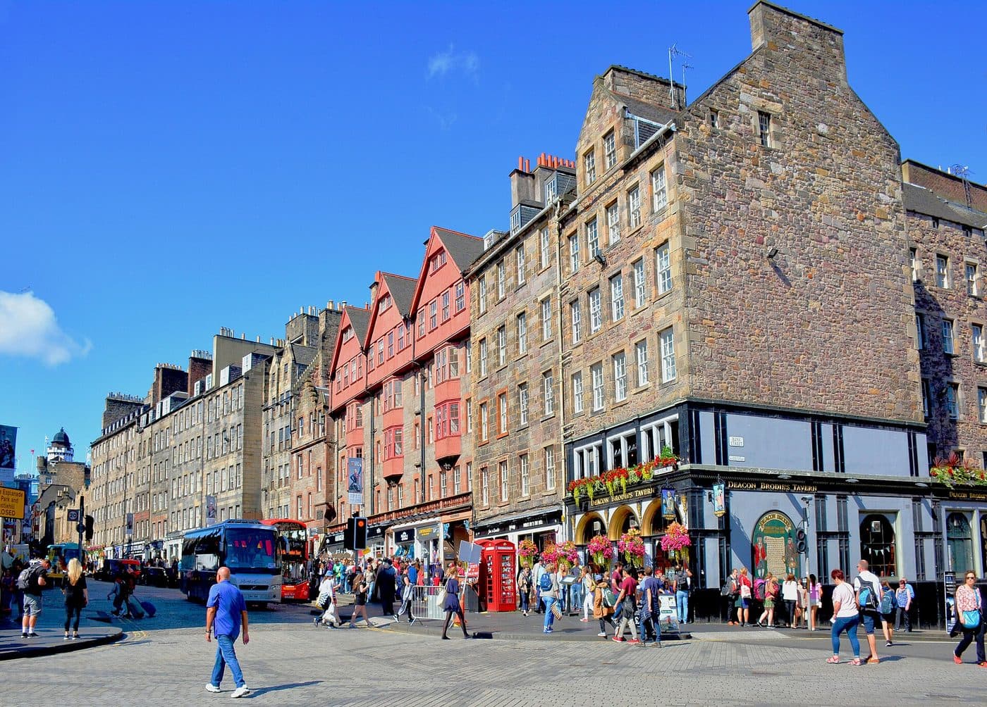 The Lawnmarket section of Edinburgh's Royal Mile on a sunny day, with Deacon Brodie's Tavern on the corner, crowds on the pavement and a tour coach on the street