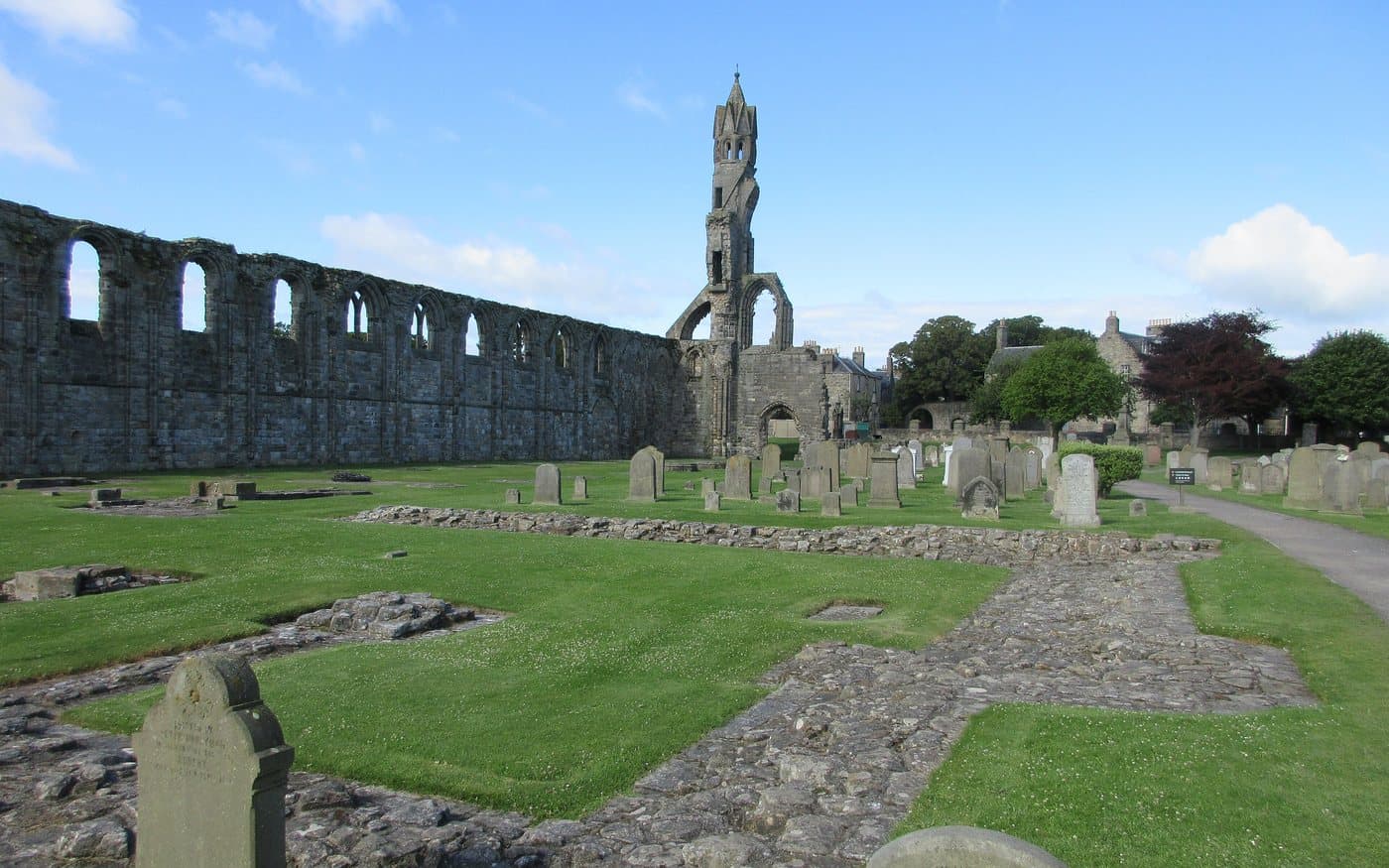 The ruined nave wall of St Andrews Cathedral with its row of empty arches, the tall gable end beyond, and a graveyard of upright headstones on mown grass